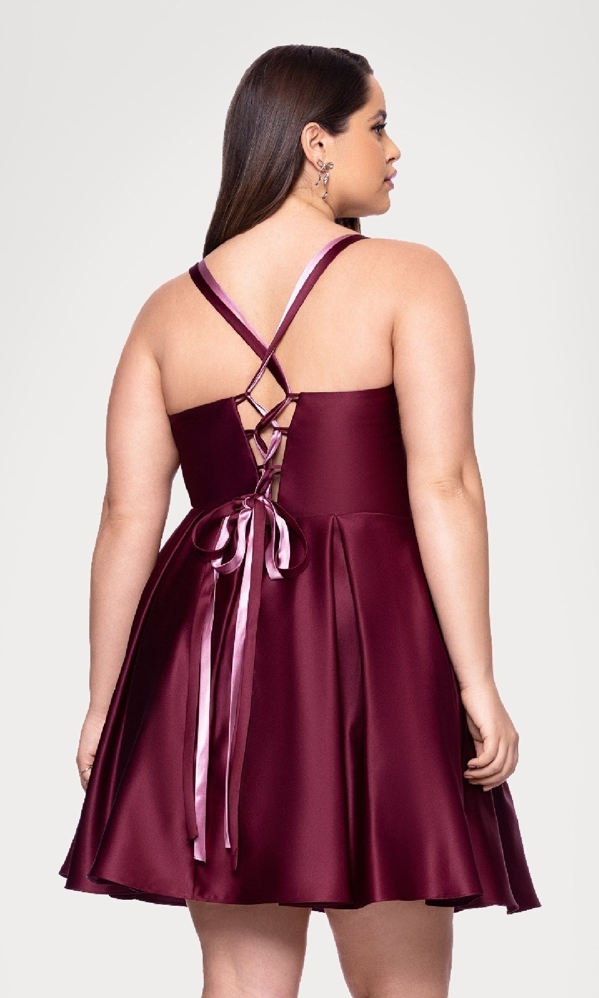 A woman models the Plus Short Wine Hoco Dress by Blondie Nites 4927BNW, featuring a sleeveless satin finish, lace-up back, and ribbon detail, set against a plain light background.